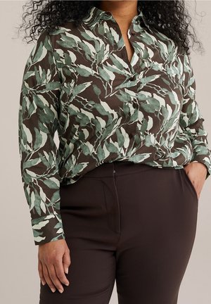 Femme portant une chemise à manches longues boutonnée avec des motifs de feuilles vertes et un pantalon sombre taille haute, une main dans la poche.