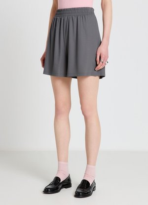 Persona che indossa pantaloncini grigi larghi, calzini rosa, mocassini neri e una maglietta rosa, in piedi su uno sfondo chiaro e uniforme.