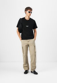 Sort t-shirt med logo, beige cargo bukser med sidelommer og sorte sneakers med hvide striber. Tilbehøret inkluderer hvide solbriller.