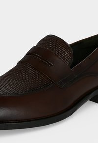 Mocassin en cuir brun foncé avec dessus à motif tissé et une bride simple sur le dessus, doté d’une semelle basse noire.