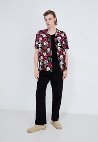 Kortærmet skjorte med sort baggrund, prydet med et blomster mønster med kranier i hvidt og rødt. Bæret med sorte bukser og brune espadrillos.