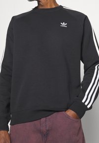 Man i svart Adidas sweatshirt med vita ränder på ärmarna och logotyp på bröstet, tillsammans med bleka, mörkröda byxor.