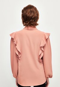 Blusa rosa con acentos de volantes en los hombros, mangas largas, cuello y una tela lisa y texturada con un corte relajado.