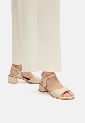 Světlé béžové sandály na blokovém podpatku s otevřenou špičkou a páskem kolem kotníku, zdobené zlatým akcentem kolem podrážky a hladkou texturou.