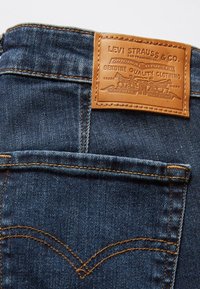 Poche arrière d'un jean Levi's bleu foncé avec un patch en cuir beige bien visible affichant le nom de la marque et le logo aux deux chevaux, cousu sur la ceinture.