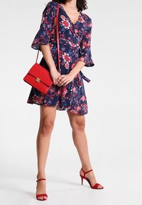 Robe portefeuille fleurie bleu marine avec manches évasées, associée à un sac bandoulière rouge et des sandales à talons rouges, mettant en valeur des motifs floraux éclatants.