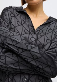 Cămașă neagră cu nasturi, realizată dintr-un material lucios cu un model complex de inimă neagră. Are guler ascuțit și mâneci lungi.