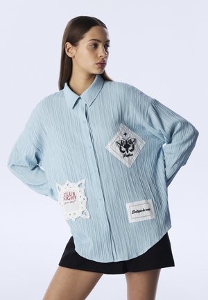 Camicia leggera azzurra, con texture, a maniche lunghe e toppe. Presenta forme a diamante, ricami neri e chiusura con bottoni. Abbinata a una gonna nera.
