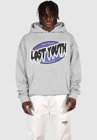 Sudadera con capucha gris que presenta un logo gráfico en negro y púrpura, con texto goteante. Combinada con pantalones cargo blancos.
