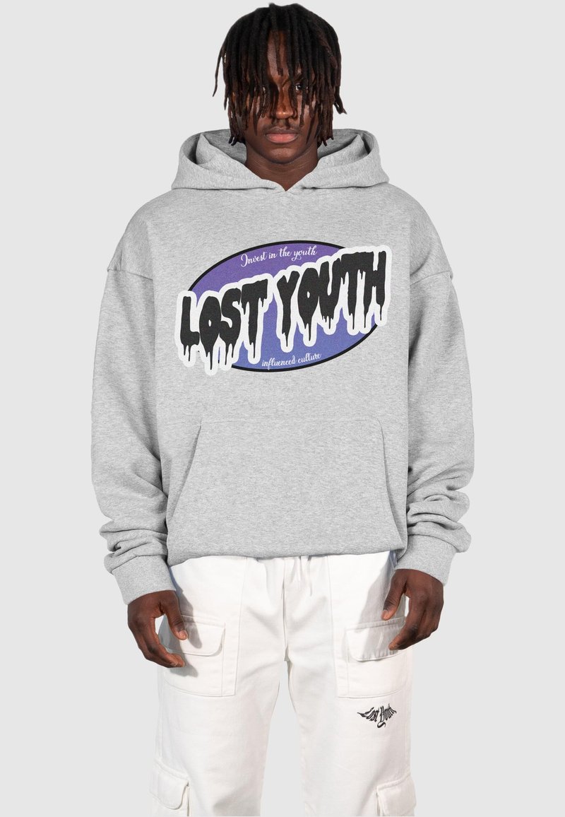 Sudadera con capucha gris que presenta un logo gráfico en negro y púrpura, con texto goteante. Combinada con pantalones cargo blancos.