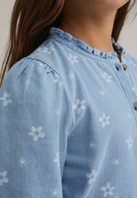 Világoskék pamut blúz fehér virágos hímzéssel és csillagmintákkal, fodros gallérral és gombos részletekkel az elején.
