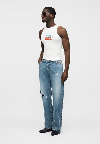Άσπρο αμάνικο τοπ με την επιγραφή "Enjoy SEX" σε μπλε και κόκκινο. Ανοιχτό μπλε τζιν με σκισίματα. Μαύρα δερμάτινα παπούτσια με μυτερή μύτη.