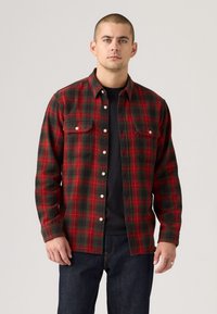 Rood en groen geruite button-up shirt met twee borstzakken en een rechte pasvorm, gedragen over een zwart t-shirt. Gemaakt van een zachte, gestructureerde stof.