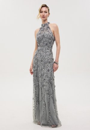 Frau trägt ein ärmelloses graues bodenlanges Kleid mit filigranen floralen Perlenverzierungen und hohem Kragen, steht vor einfarbigem Hintergrund.