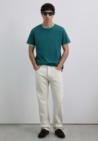 Tricou scurt cu mâneci, de culoare teal, cu margini albe, asortat cu pantaloni bej deschis și pantofi maro închis cu șireturi. Modelul stă în picioare cu mâinile în buzunare.
