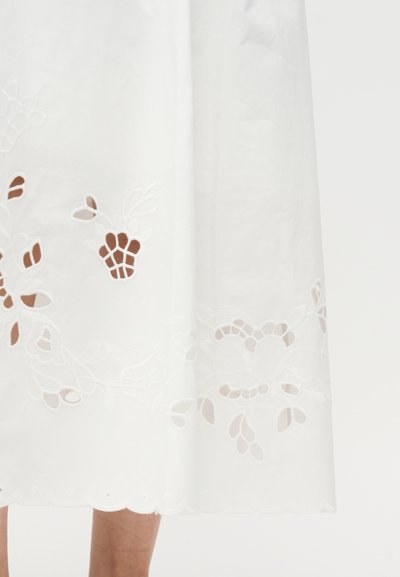 Hvid midi nederdel med blomsterudskåret broderi og bølget kant, båret af en person, der står mod en ensfarvet baggrund.