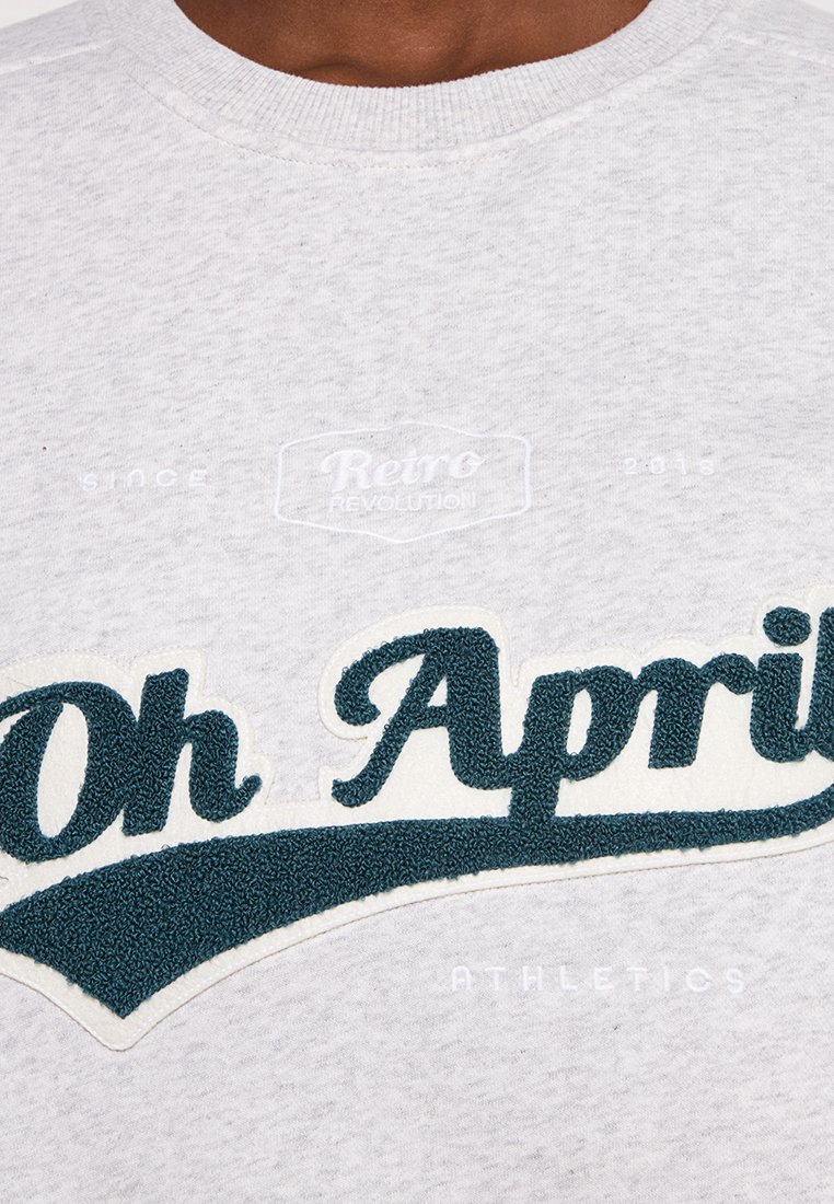 Hellgrau Sweatshirt mit strukturierten dunkelblauen gestickten Buchstaben "Oh April" und "ATHLETICS" sowie einer glatten Stoffoberfläche.