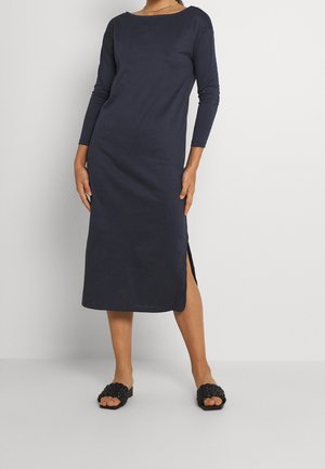 Granatowa sukienka z długim rękawem wykonana z miękkiego materiału, z bocznymi rozcięciami i dekoltem w łódkę. Noszona z czarnymi plecionymi sandałami typu slide.