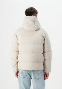 Beige puffersdzseki, illeszkedő kapucnival, tűzött mintával és rugalmas mandzsettákkal; világoskék farmernadrág felett viselve, mutatva a laza, meleg stílust.