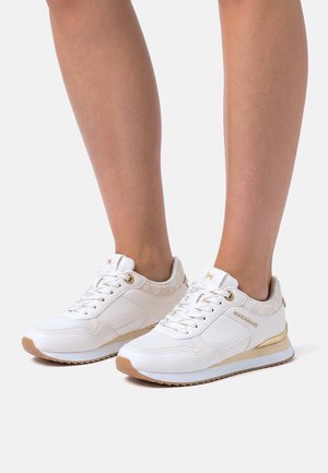 Witte lage sneakers met gouden accenten en textuurdetails, gedragen op blote benen tegen een effen achtergrond.
