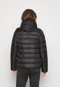 Person som bär en svart puffjacka med huva, står med ryggen mot kameran framför en enkel vit bakgrund.