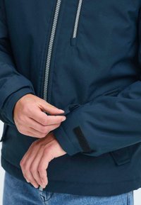 Persona che regola la fascia in Velcro sul polsino di una giacca blu navy con zip argentata e jeans di denim.