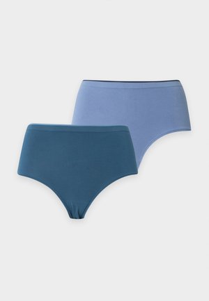 Femme debout pieds nus portant un soutien-gorge gris clair et un sous-vêtement taille haute bleu sarcelle foncé sur un fond clair uni.