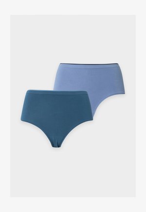 Femme debout pieds nus portant un soutien-gorge gris clair et un sous-vêtement taille haute bleu sarcelle foncé sur un fond clair uni.