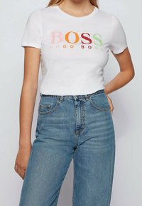 Camiseta blanca de manga corta con la palabra "BOSS" en letras multicolores y "HUGO BOSS" en texto más pequeño debajo. Combinada con unos jeans de mezclilla azul de tiro alto.