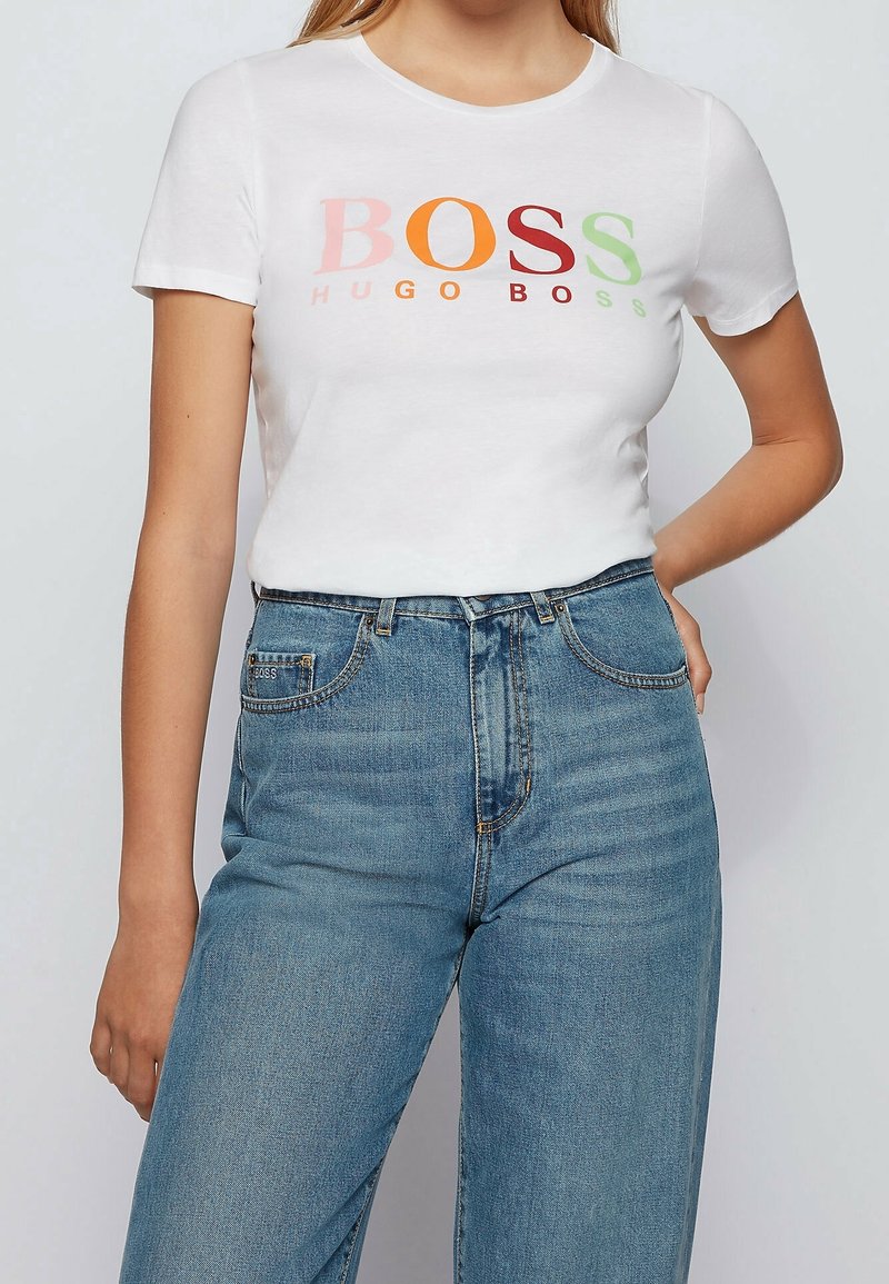 Camiseta blanca de manga corta con la palabra "BOSS" en letras multicolores y "HUGO BOSS" en texto más pequeño debajo. Combinada con unos jeans de mezclilla azul de tiro alto.
