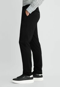 Schwarze maßgeschneiderte Hose mit entspannter Passform, seitlichen Taschen und glatter Textur. Kombiniert mit schwarz-weißen Sneakers mit einem Logo.