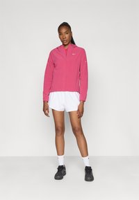 Veste légère rose avec capuche, fermeture éclair et logo Nike discret. Associée à un short blanc et des chaussures de sport noires. Texture lisse.