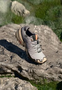 Hellegraue Trailschuhe mit schwarzen und orangenen Akzenten, ausgestattet mit einem strukturierten Obermaterial, Schnürverschluss und robustem Sohlen-Design.