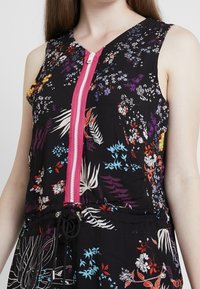 Blommig jumpsuit i svart tyg med en rosa dragkedja, ärmlös design och justerbar midja med dragsko; livfulla flerfärgade blommönster.