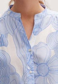 Chemise à manches longues bleu clair avec un motif floral dans différentes nuances de bleu. Le tissu semble lisse avec un léger éclat, doté d'un col mandarin.