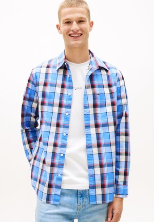 Giovane uomo sorridente, che indossa una camicia a quadri blu, bianca, nera e rossa sopra una T-shirt bianca e jeans azzurri, in piedi davanti a uno sfondo bianco.