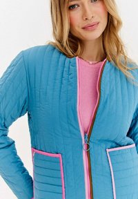 Pikowana niebieska kurtka z różowym zamkiem i wykończeniem, z dwoma przednimi kieszeniami. Materiał ma przeszyte linie dla uzyskania faktury i ciepła.