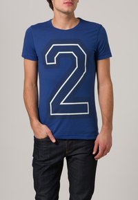 Blå kortärmad t-shirt med ett stort vitt konturliknande nummer "2" i mitten, tillverkad av mjukt tyg, i kombination med mörk jeans.