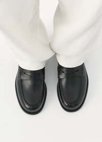 Mustad nahast loafers, millel on sile tekstuur, eristuva õmblusega disain ja dekoratiivne rihm jala peal. Osaliselt nähtavad valged püksid.