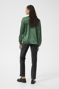 Grüne Satinsbluse mit langen Ärmeln und gerafften Manschetten, kombiniert mit hoch taillierten schwarzen Jeans und schwarzen Slippern. Glatte Textur.