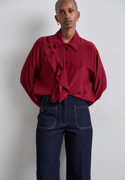 Μαρό μ blouse με λεπτομέρεια από φλόντζες στον ένα ώμο, κουμπότρυπα μπροστά και φαρδιές μανσέτες. Συνδυάζεται με ψηλόμεσα σκούρα τζιν παντελόνια.