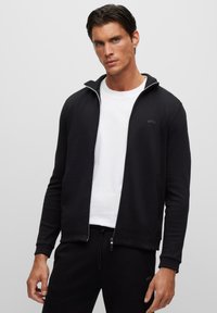 Svart zip-up-jacka med figursydd design, utrustad med ett vitt innerfoder och BOSS-logotyp på bröstet, kombinerad med en vit T-shirt under.