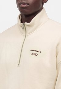 Homme portant un sweat crème à fermeture éclair quart de zip avec le logo "Abercrombie & Fitch" sur la poitrine, sur un fond uni.