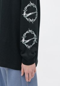 Rukáv čiernej dlhoprstej košele s dvoma striebornými kruhmi z ostnatého drôtu, zobrazujúcimi štýlové Nike swooshy, oblečený osobou v svetlomodrých nohaviciach.