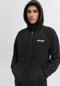 Zwarte zip-up hoodie met geribbelde accenten, kangoeroezakken en een klein wit logo op de borst. Gemaakt van zacht, textuurmateriaal. Inclusief capuchon.