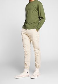 Hombre de pie con pantalones cargo beige, zapatillas altas blancas y una camiseta de manga larga verde con un logo en el pecho, con las manos en los bolsillos sobre un fondo gris.