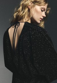 Frau mit welligem blondem Haar, die ein schwarzes, glitzerndes Kleid mit offenem Rücken trägt, das von dünnen Trägern gehalten wird, vor einem grauen Hintergrund.