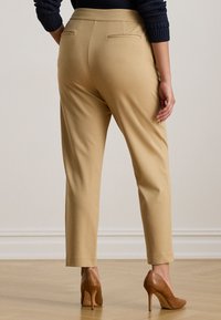 Moteris vilkinti smėlio spalvos pritaikytus kelnes, rudas aukštakulnius batus ir tamsią ilgąsleeves viršutinę dalį, stovinti ant šviesaus medinio grindų.