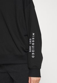 Sweat-shirt noir à coupe décontractée, avec inscription blanche brodée "MISSGUIDED EST 2009" sur la manche gauche. Matière en coton doux.