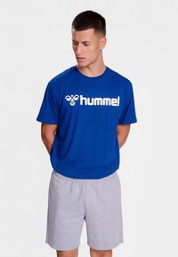 Blå bomullst-shirt med vitt "hummel"-logo, kombinerad med ljusgrå shorts. Enkel design, kortärmad, normal passform.